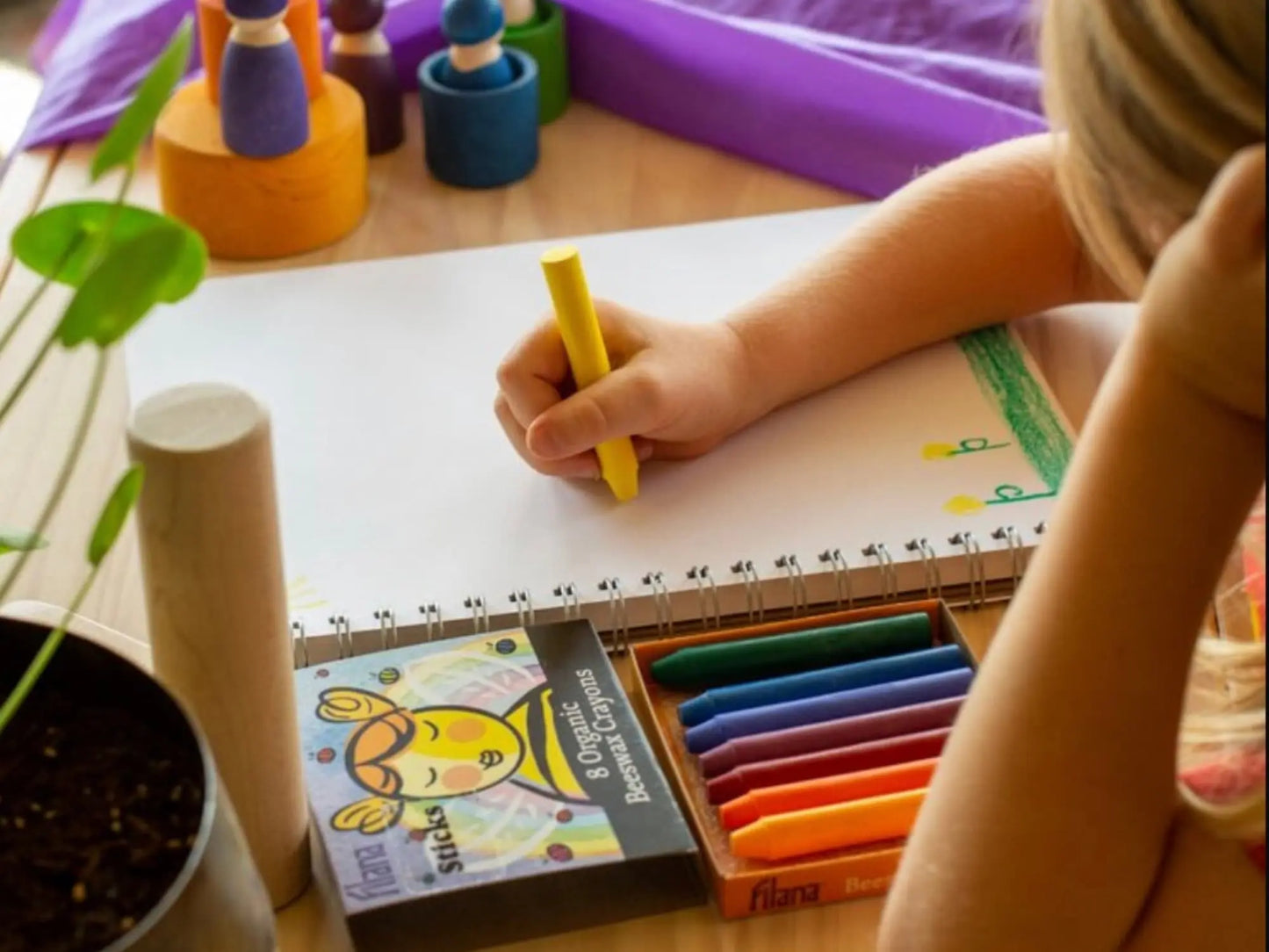 Child drawing with a yellow Filana beeswax stick crayon on a notebook with art supplies around