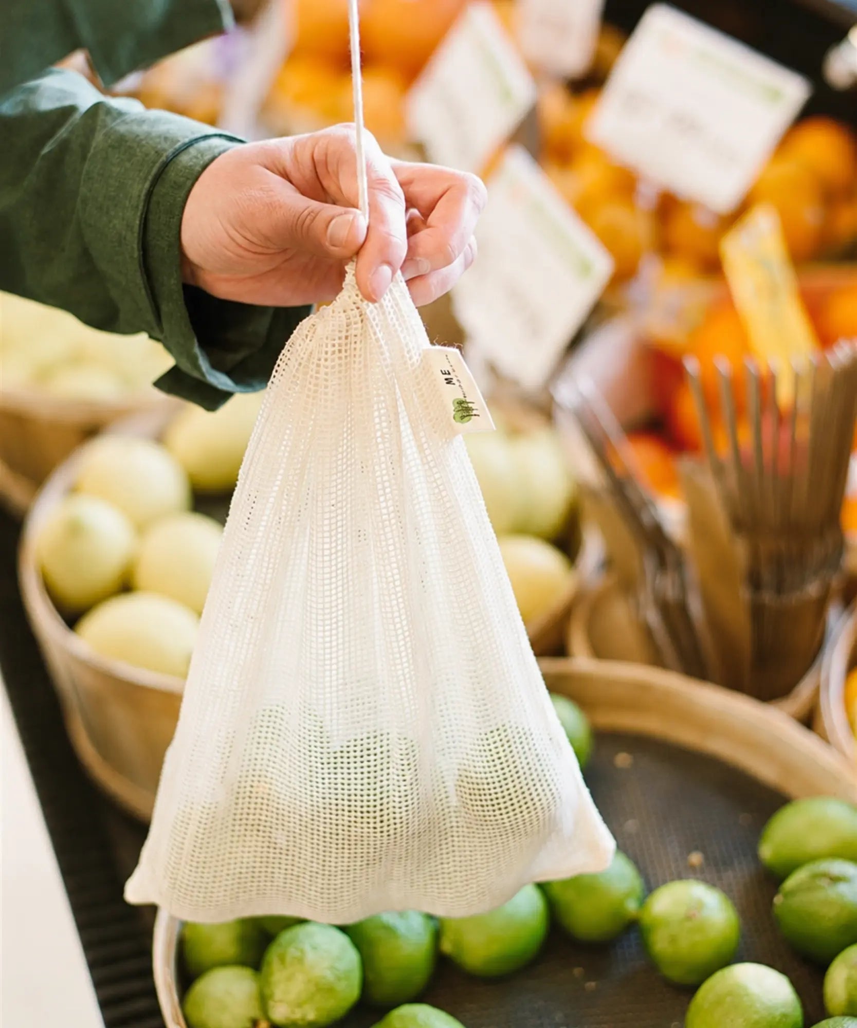 Hand holding an organic cotton mesh produce bag filled with fresh produce while shopping at a farmers market.