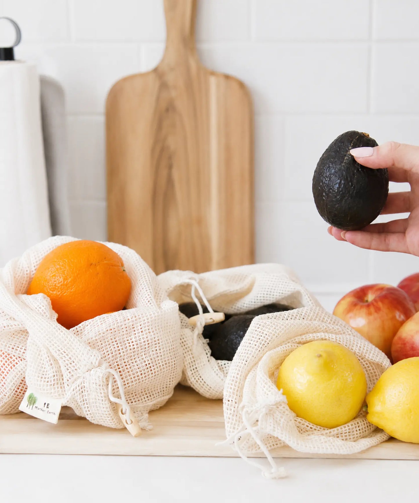 Hand holding an avocado over organic cotton mesh produce bags filled with fresh produce