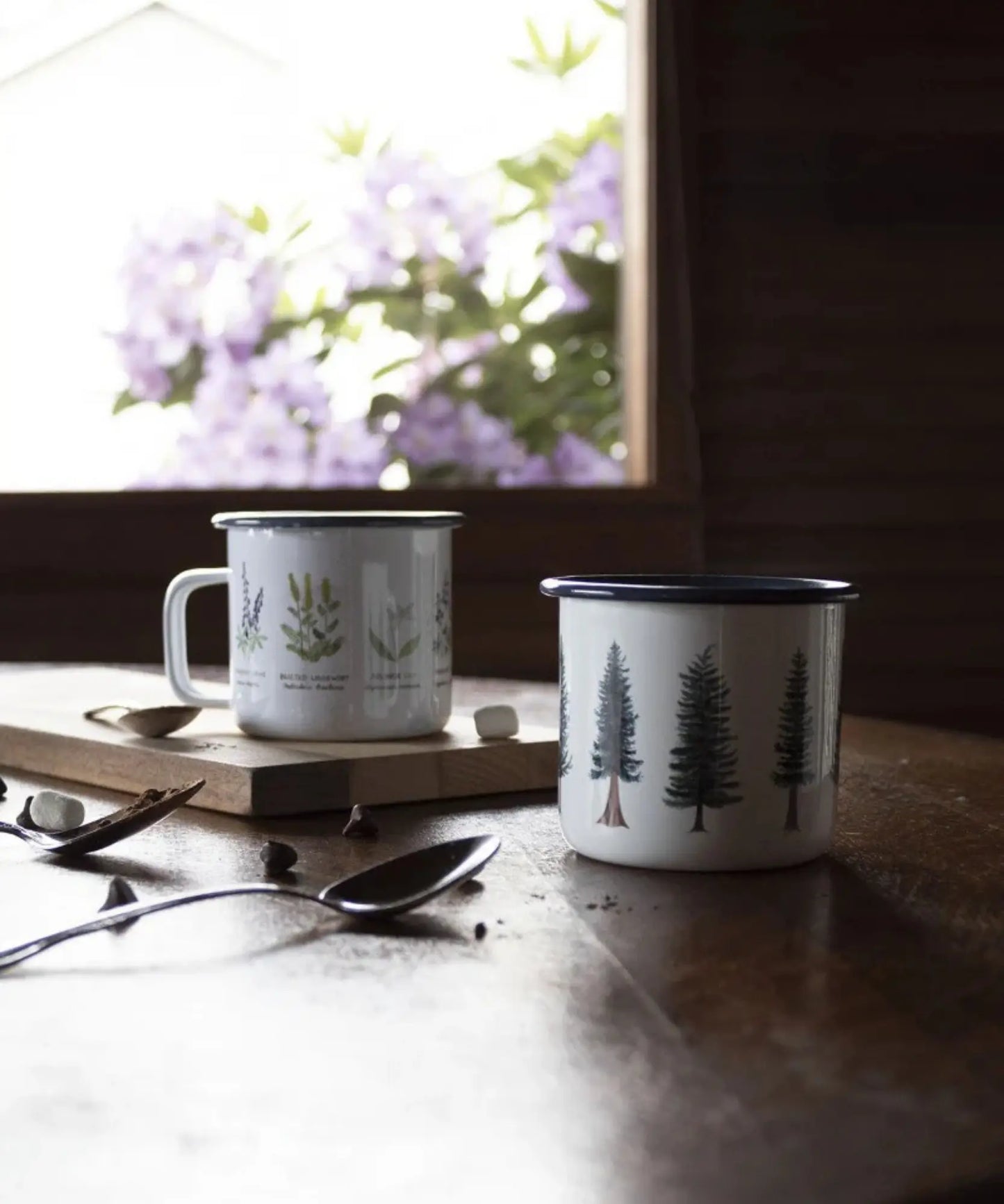 Two mugs on a wooden surface with a window in the background showing purple flowers.
