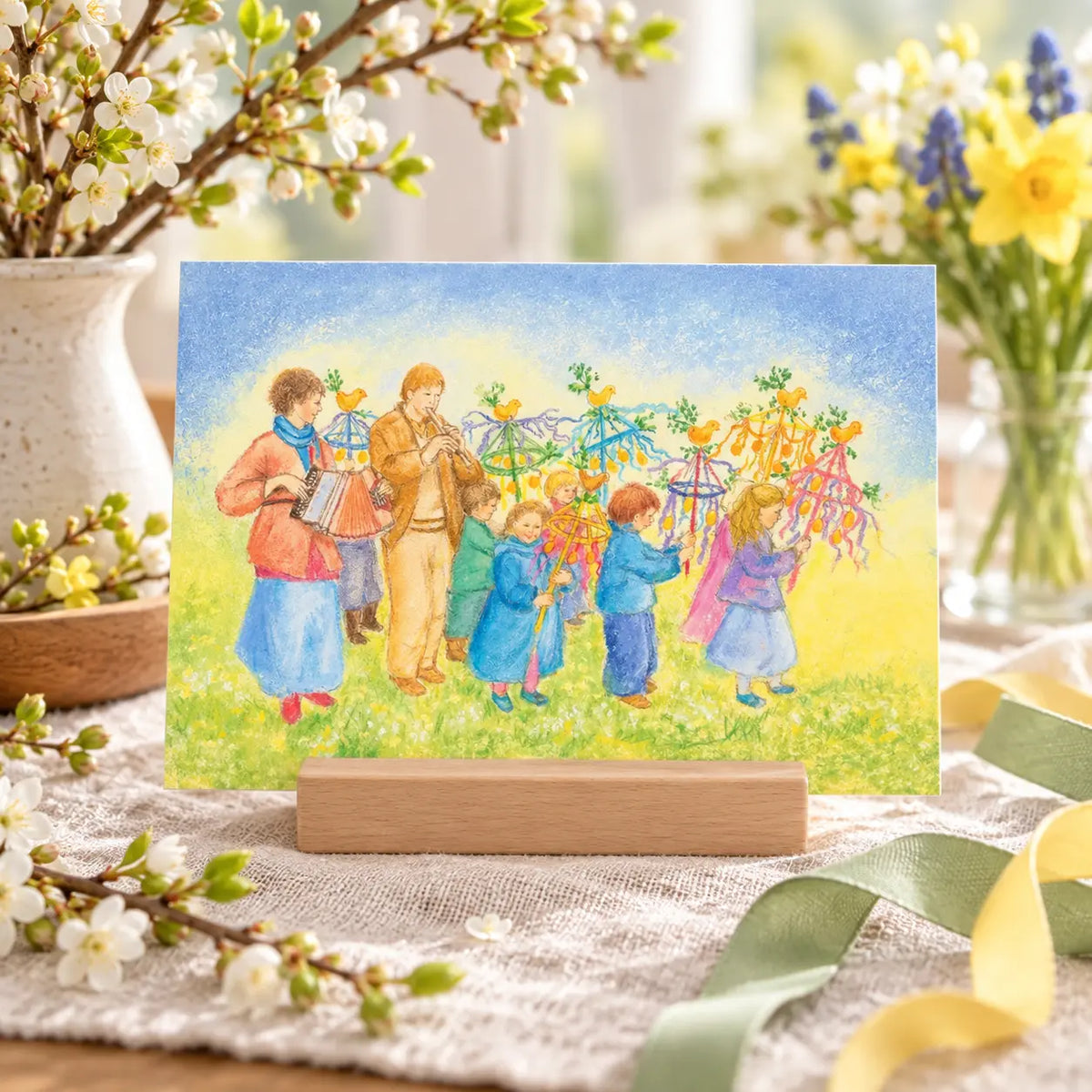 Palm Sunday Procession postcard by Marjan van Zeyl displayed in a wooden holder on a spring nature table with blossoming branches, yellow daffodils, and soft green ribbons.