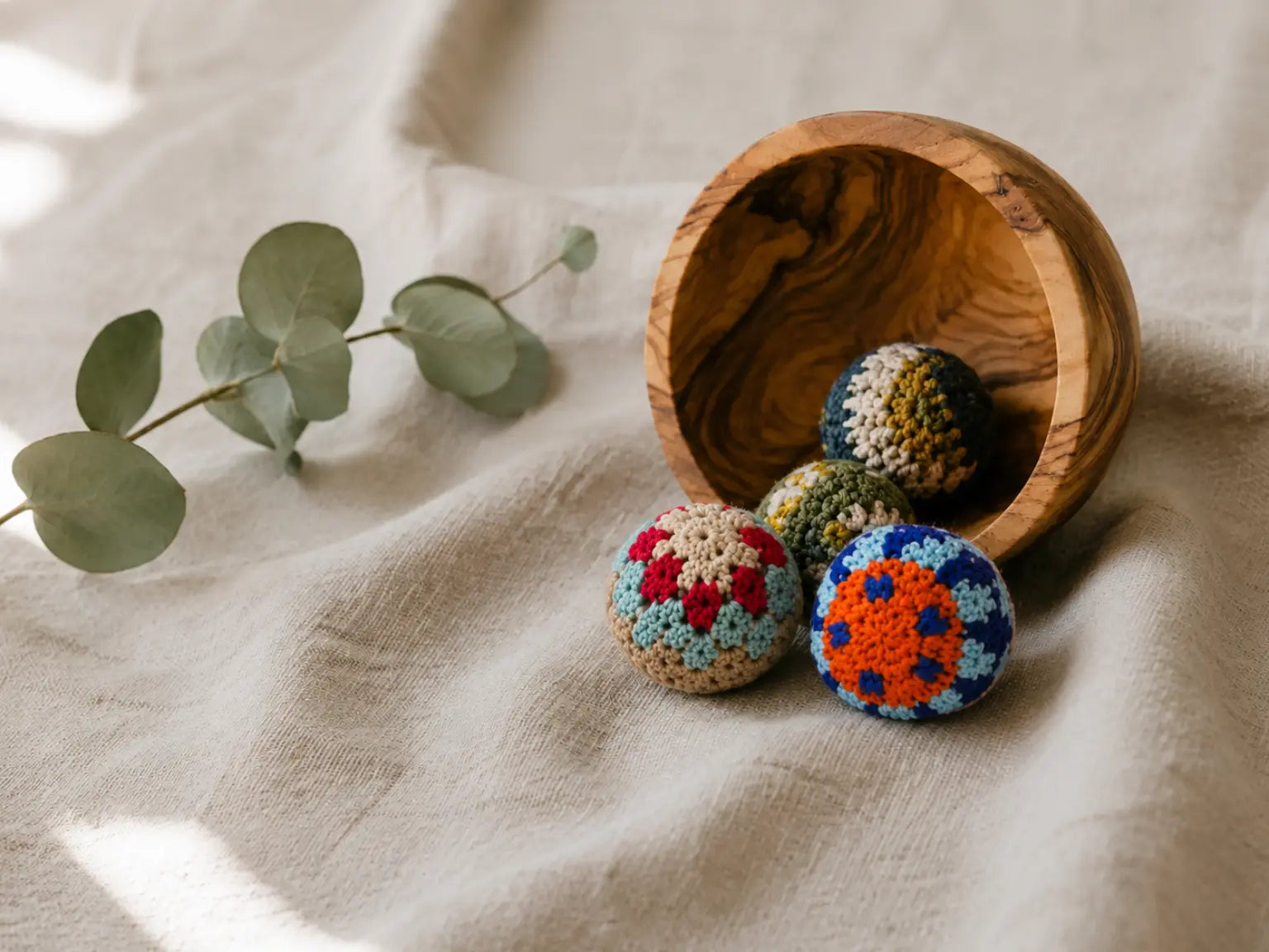 Four crocheted Fair Trade Pelotie cotton balls arranged next to an olive wood bowl on a beige linen fabric