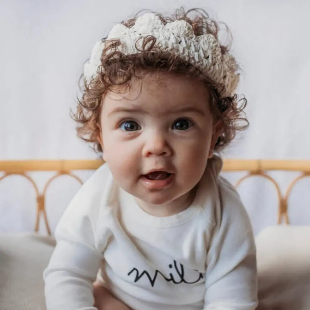 Baby wearing a white onesie with text and a knitted hat, sitting on a couch.