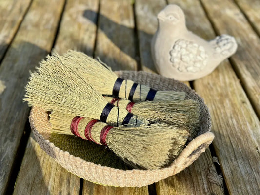 Two mini whisk brooms with cord wrapped handles in a woven basket on a wooden surface