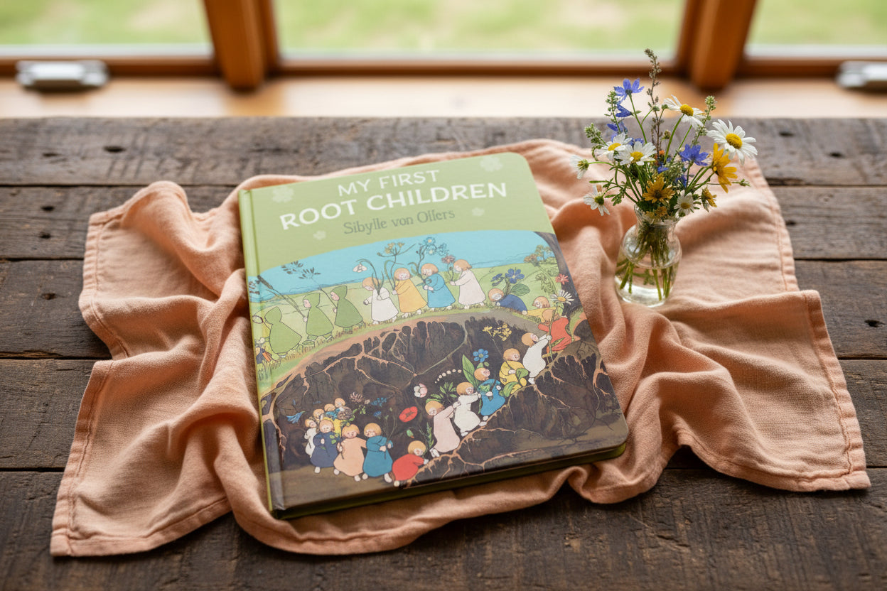 Children's book titled 'My First Root Children' on a wooden surface with a pink cloth and flowers.