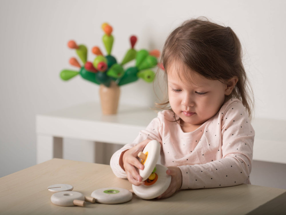 Child playing with wooden lacing toy on a table with a toy cactus plant in the background
