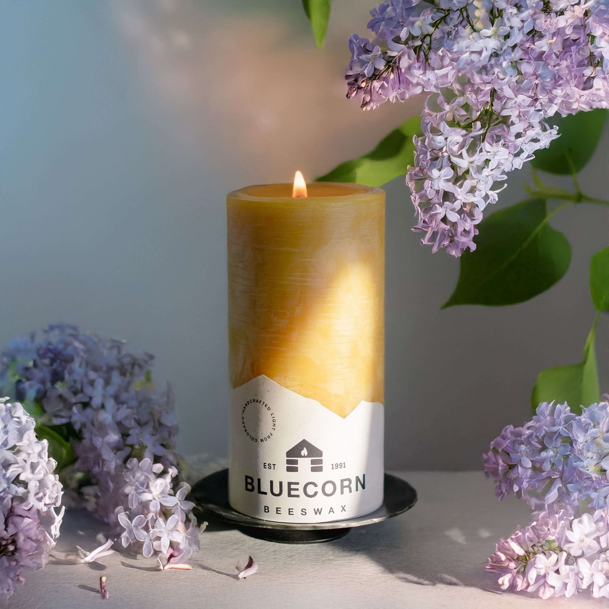 Candle with Bluecorn Beeswax label surrounded by lilac flowers