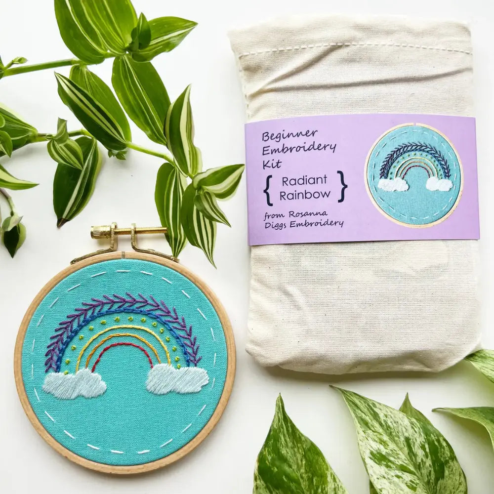 Radiant Rainbow beginner embroidery kit with completed rainbow embroidery hoop beside project bag and green houseplants