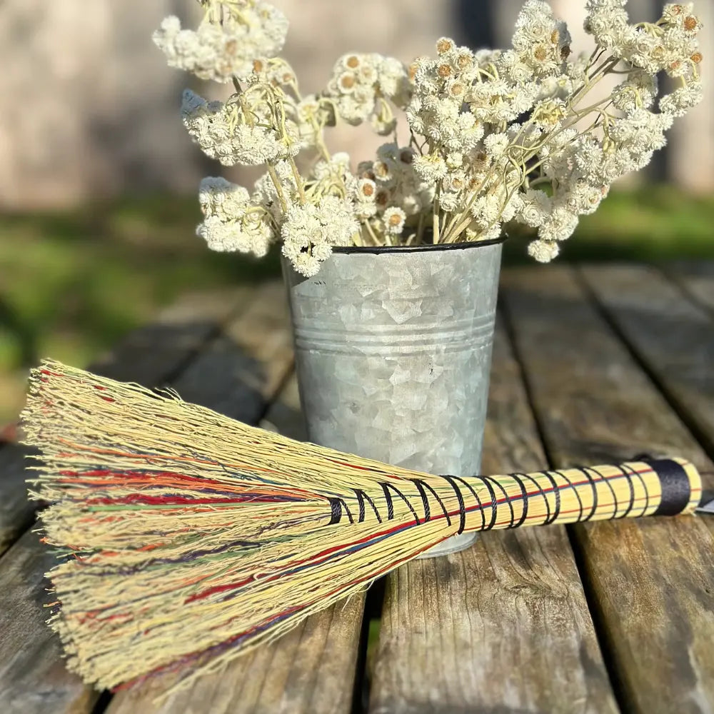 Broom with decorative handles next to a metal bucket filled with white flowers on a wooden surface.
