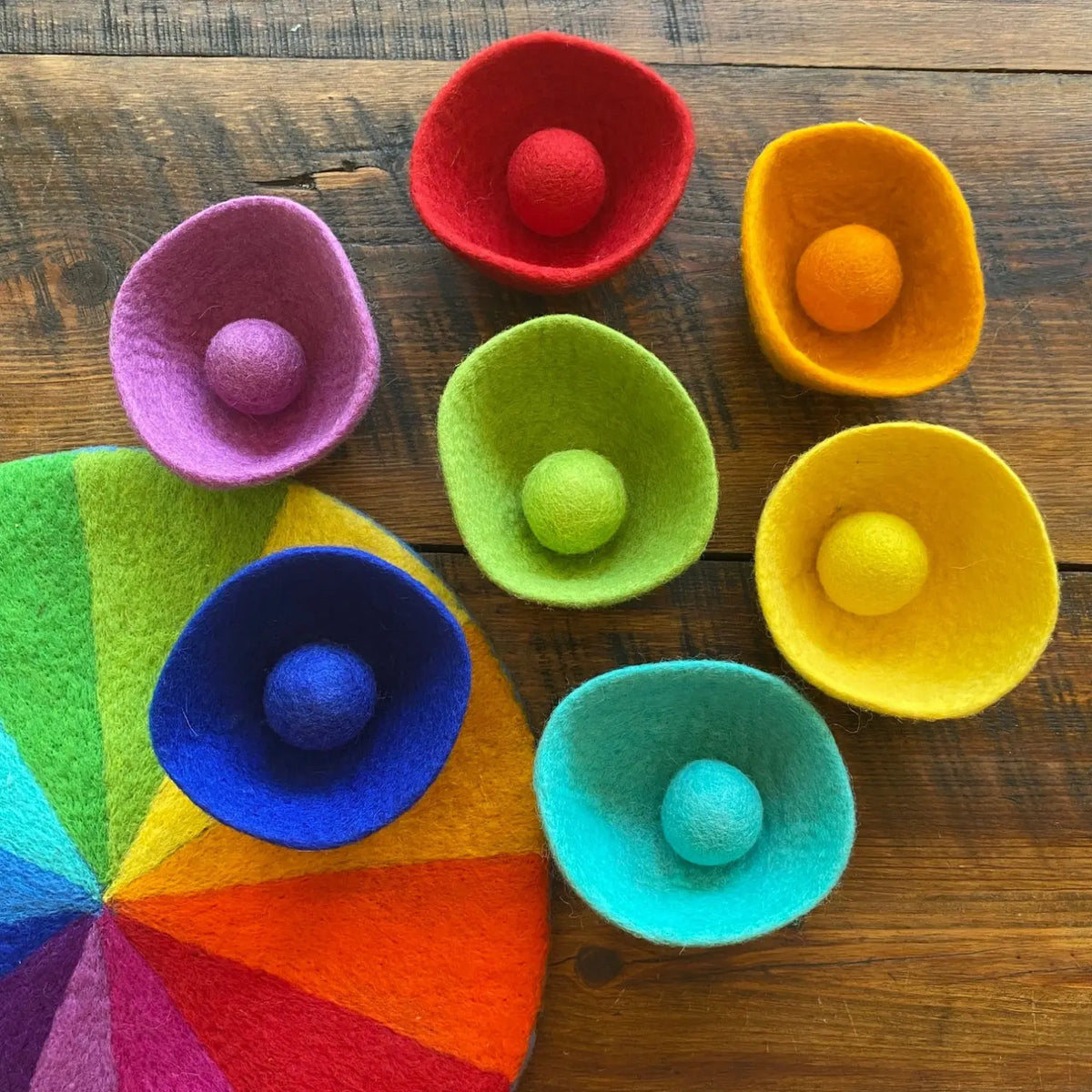 Seven rainbow wool felt bowls holding matching felt balls, arranged on a 12-color felt play mat.