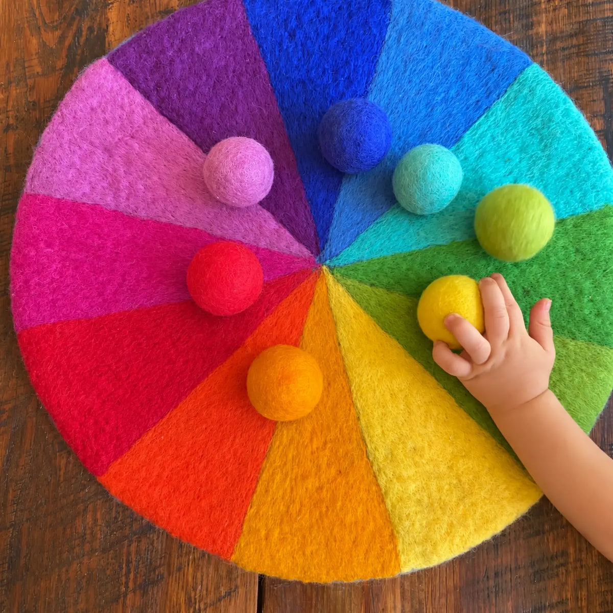 Child’s hand places a yellow felt ball on a 12-color rainbow wool play mat