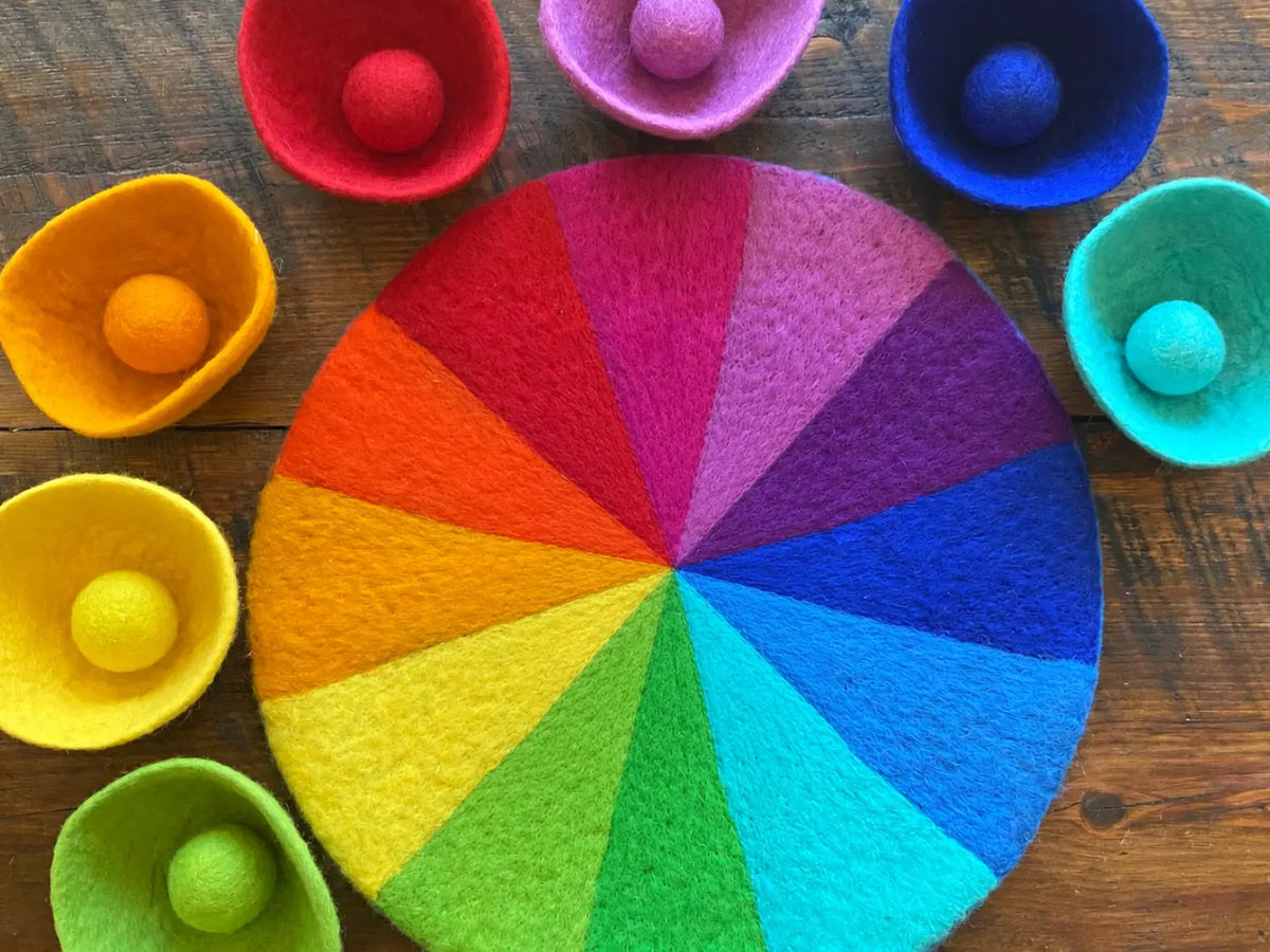 Seven colorful felt bowls arranged around a rainbow felt mat and storage bag; mat sold separately.
