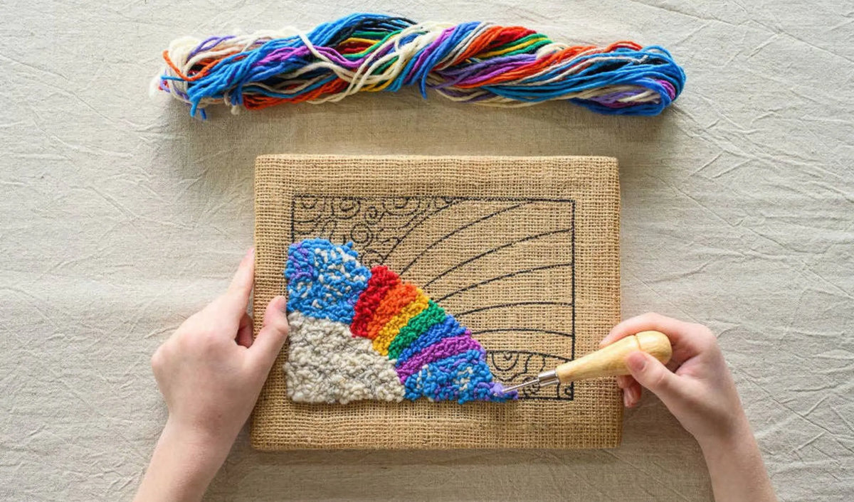 Overhead view of hands hooking a wool rainbow and blue sky on a burlap frame, with rainbow yarn nearby