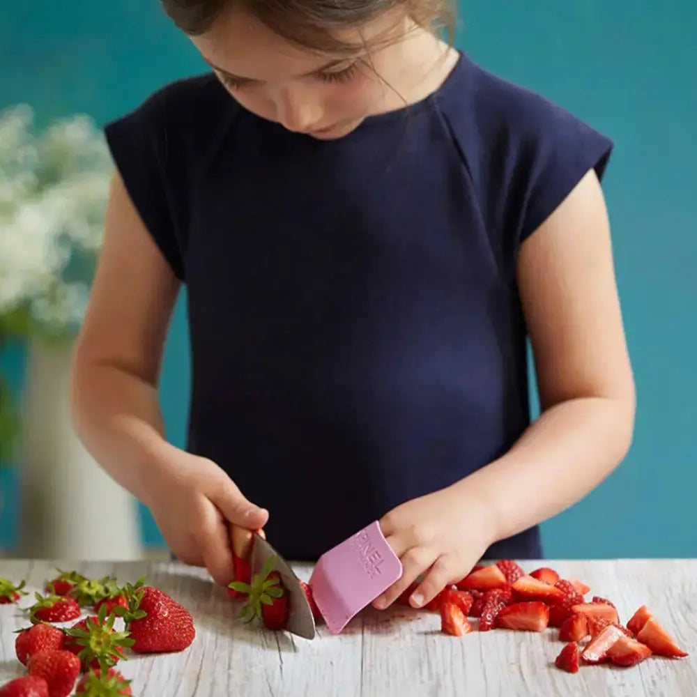 Child cutting strawberries with a pink kitchen knife on a wooden table.