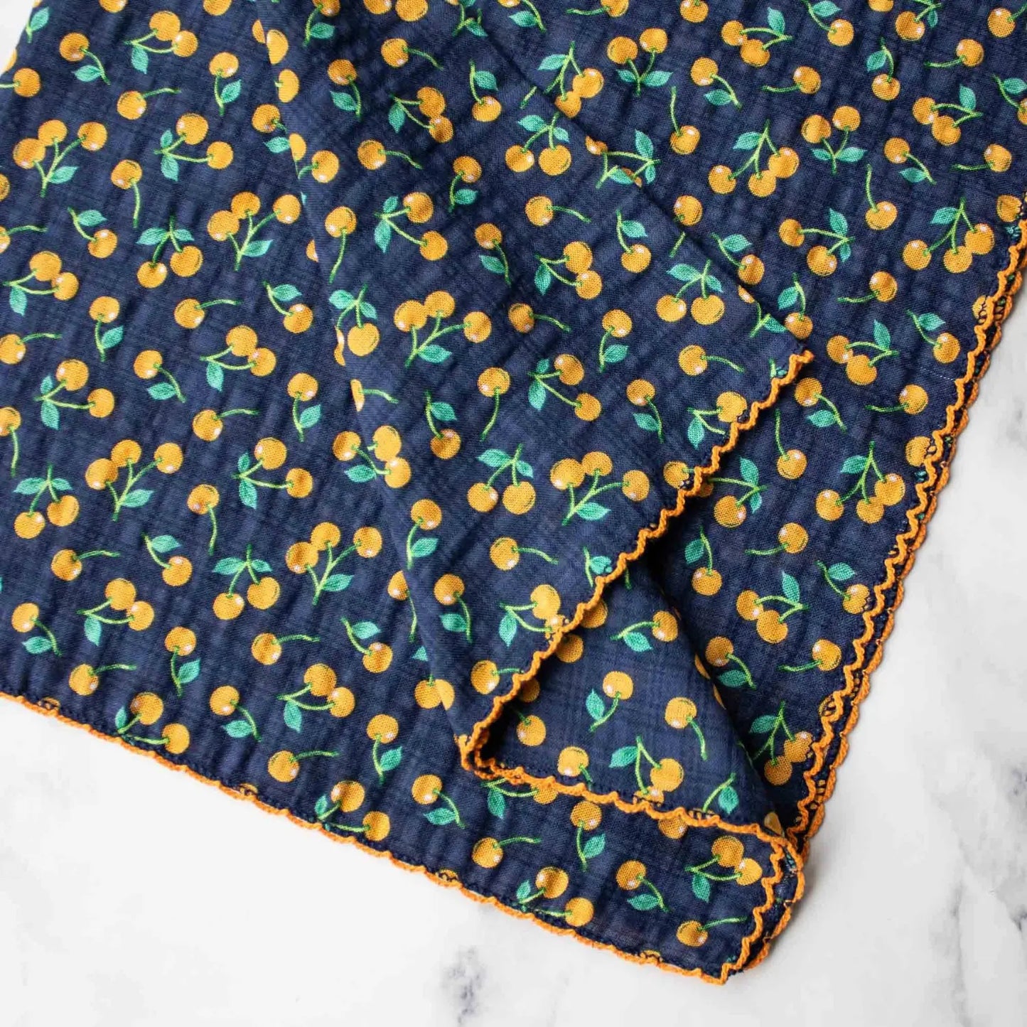 Golden Cherry reusable lunch napkin on navy blue with a small golden cherry print and orange decorative edging close up detail