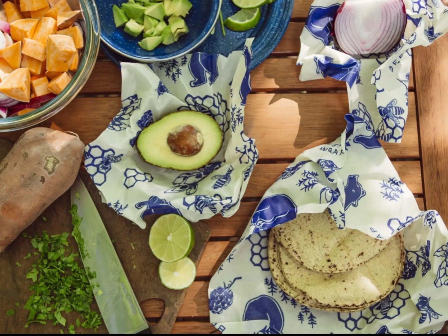 Foods wrapped in blue and white patterned beeswax wraps on a wooden surface with ingredients like avocados, tortillas, and sweet potatoes.