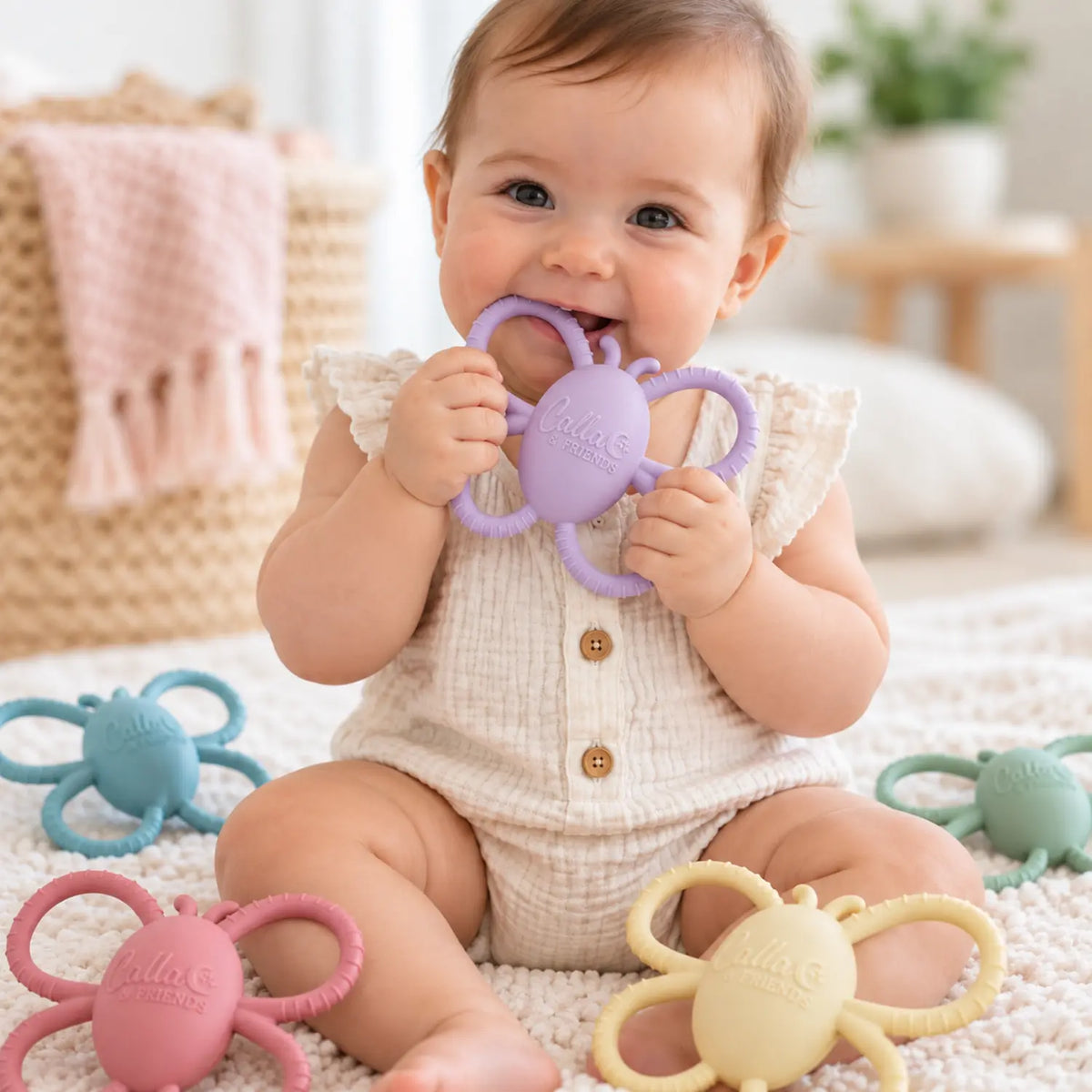 Baby holding and chewing a lilac silicone butterfly rattle teether with pastel colors nearby