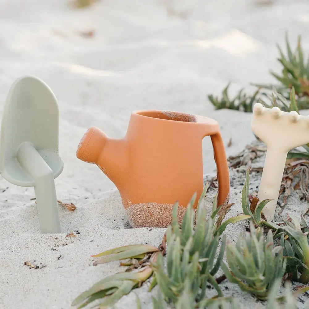 Children's silicone gardening tools including a watering can, shovel, and trowel on sandy ground with plants.