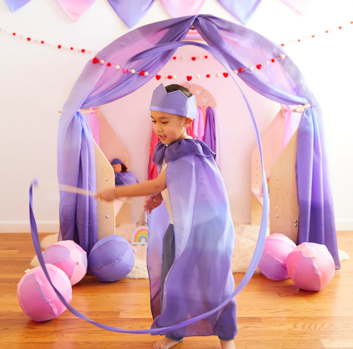 Child wearing butterfly colorway cape, crown, and streamer playing in front of a Waldorf play arch