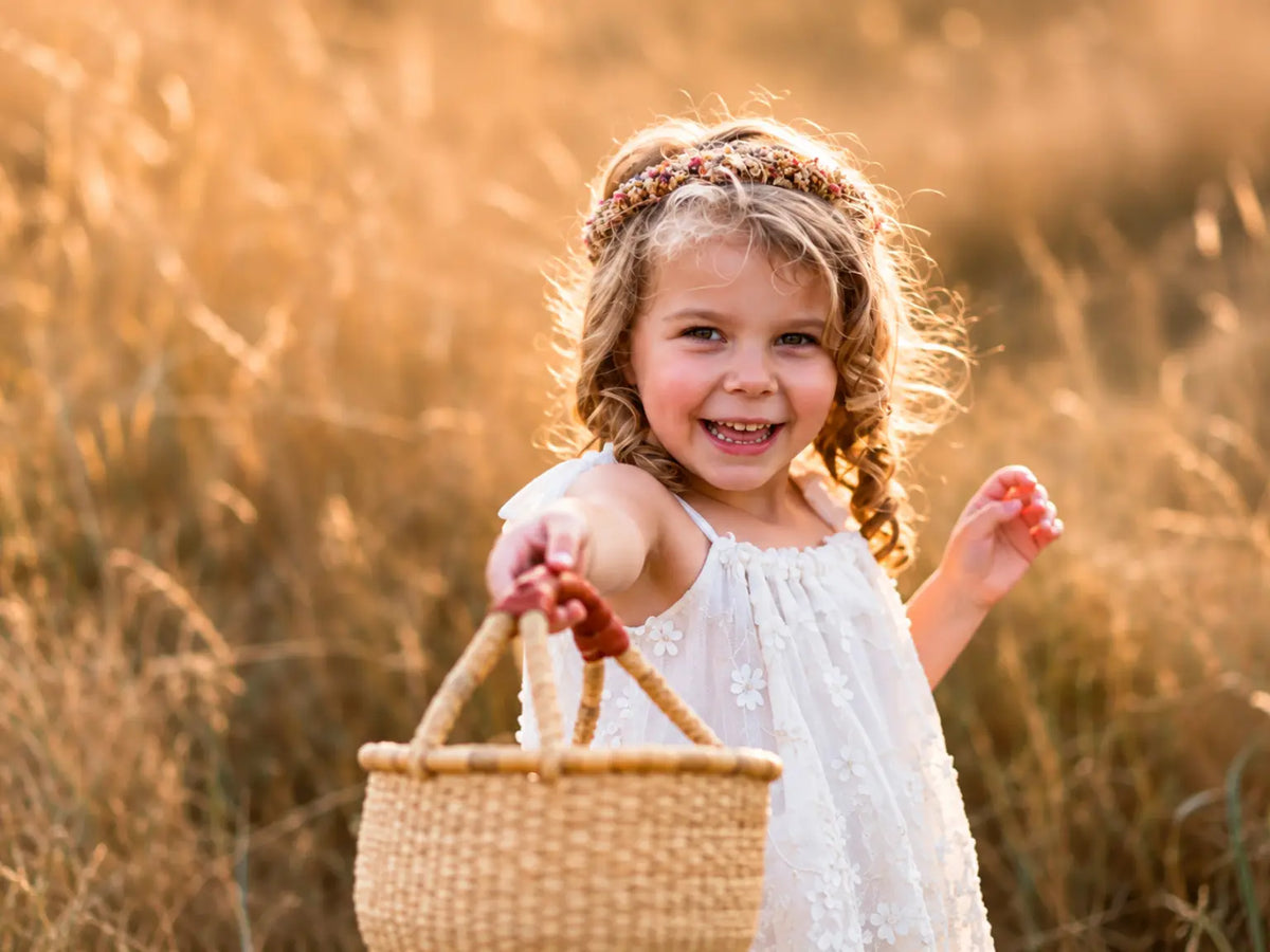 Young girl in a white embroidered dress smiling as she holds out a small handwoven Bolga gathering basket in a sunlit field.