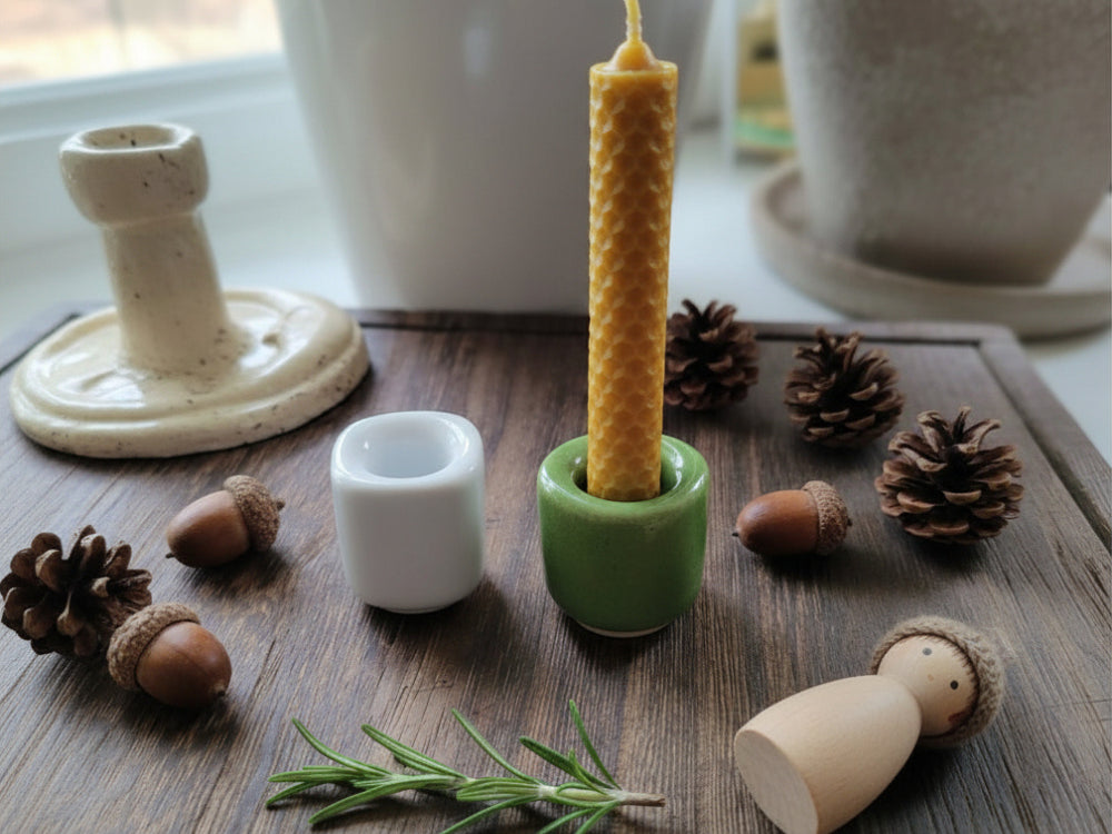 Decorative items including a beeswax candle, small white candle holder, green holder, wooden figurine, pinecones, and acorns on a wooden surface.