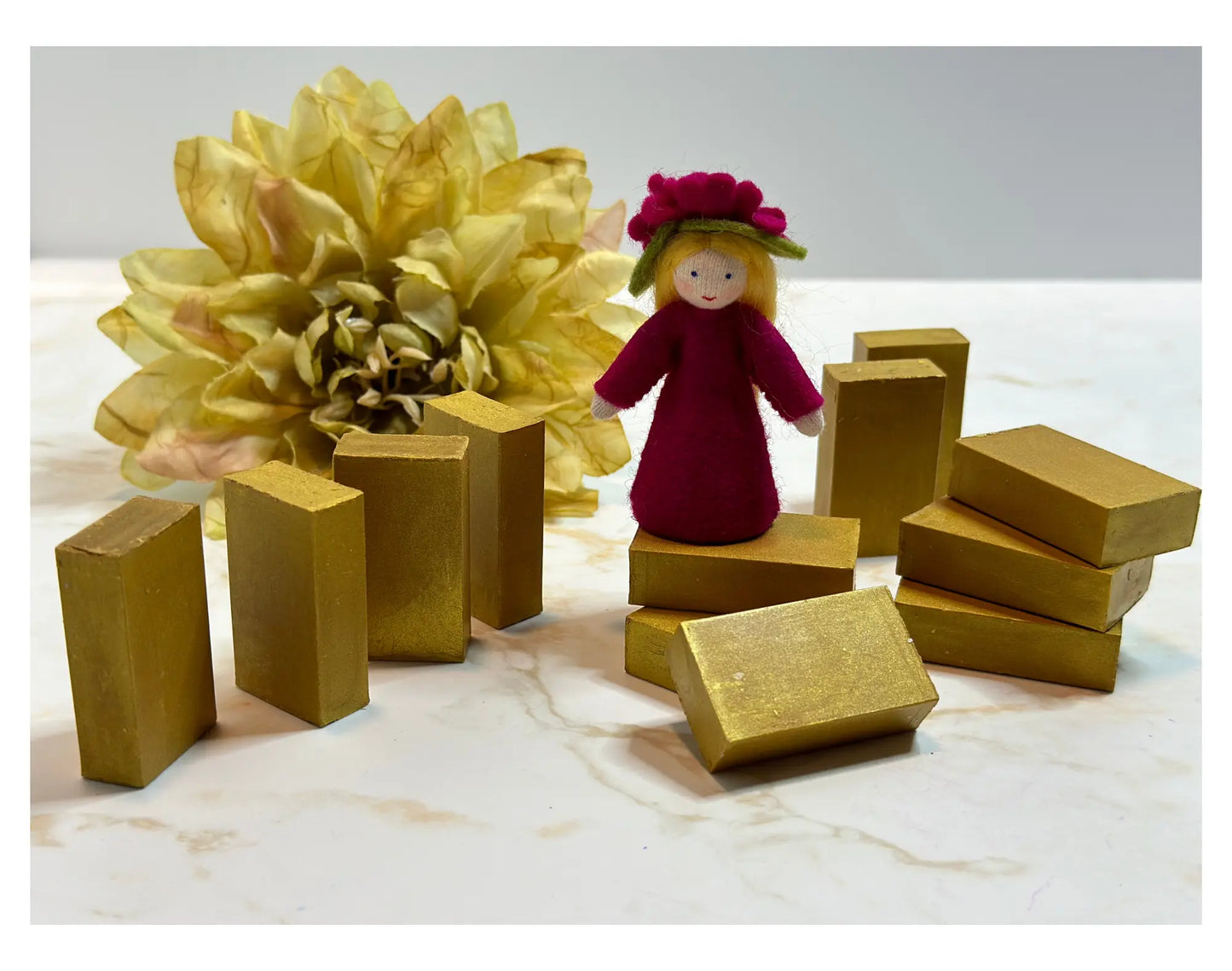 Gold Stockmar Block Crayons and a small figurine with a magenta dress and flower headband on a light background