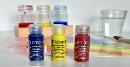 Set of Stockmar watercolor bottles on a white surface with a glass of water and paintbrushes in the background.