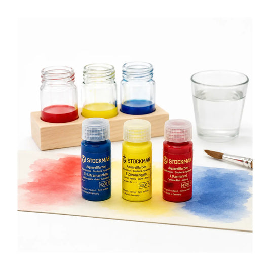 Three Stockmar watercolor paint bottles in carmine red, lemon yellow, and ultramarine blue with paint jars, brush, and wet-on-wet color washes.