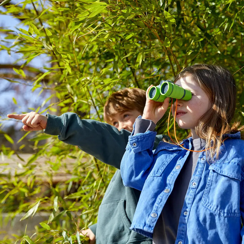 Two children outdoors, one using binoculars and pointing, with greenery in the background.