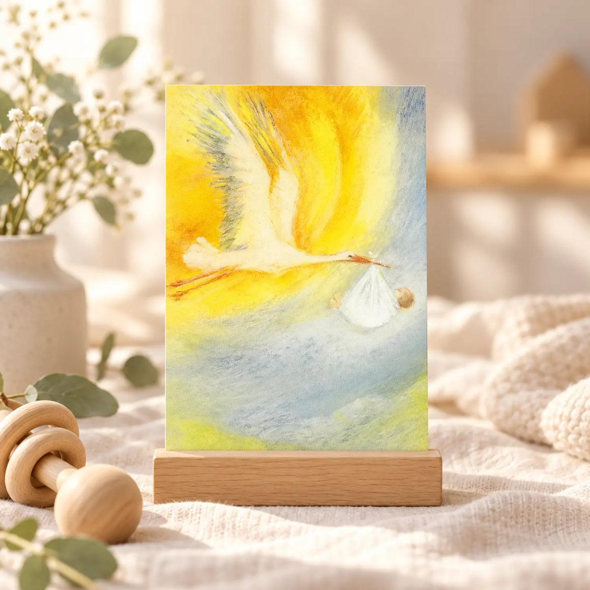 The Stork postcard by Marjan van Zeyl displayed in a wooden holder on soft natural linen with a wooden baby rattle, eucalyptus stems, and a cozy nursery setting.