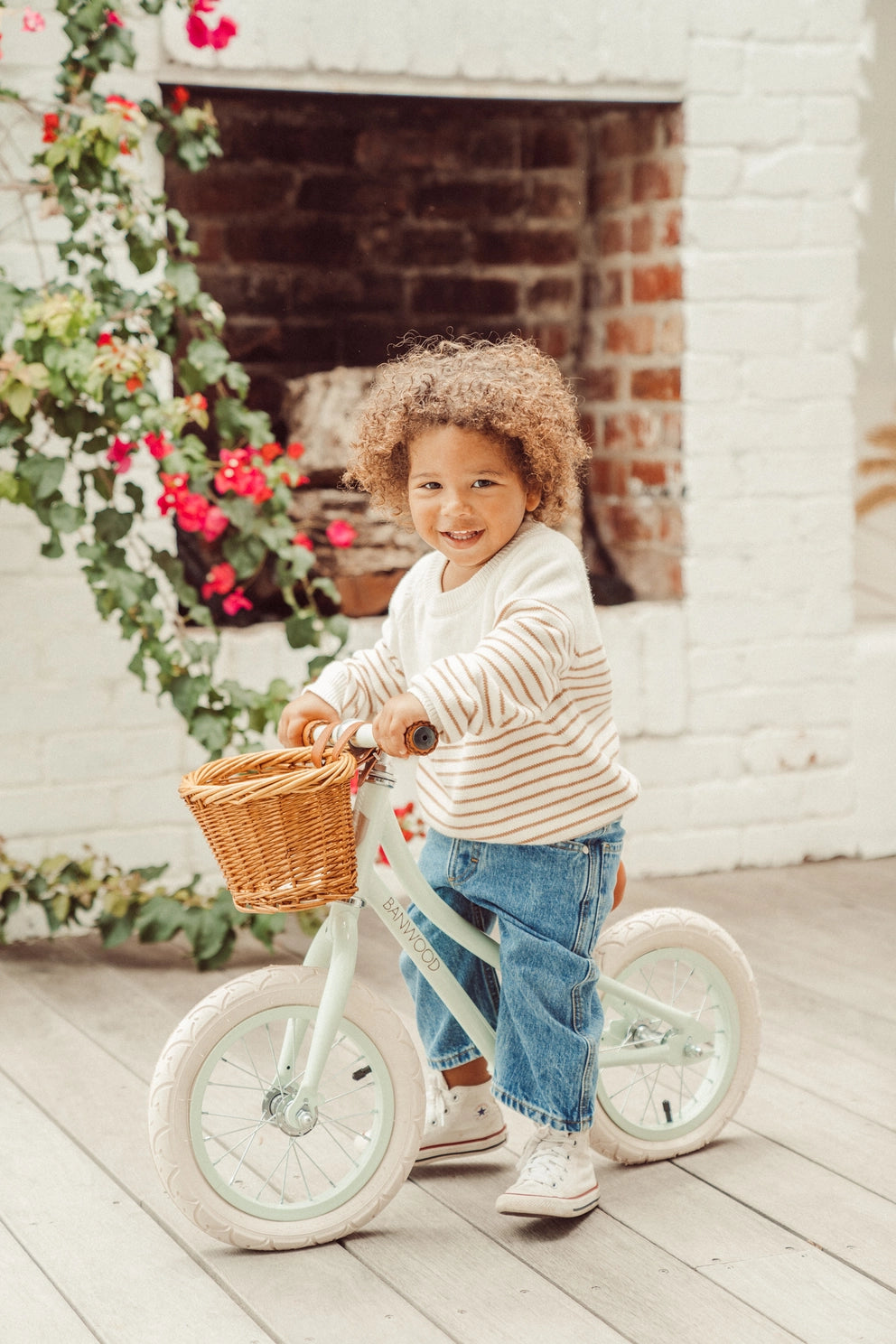Child riding a balance bike with a basket in front of a white brick wall.