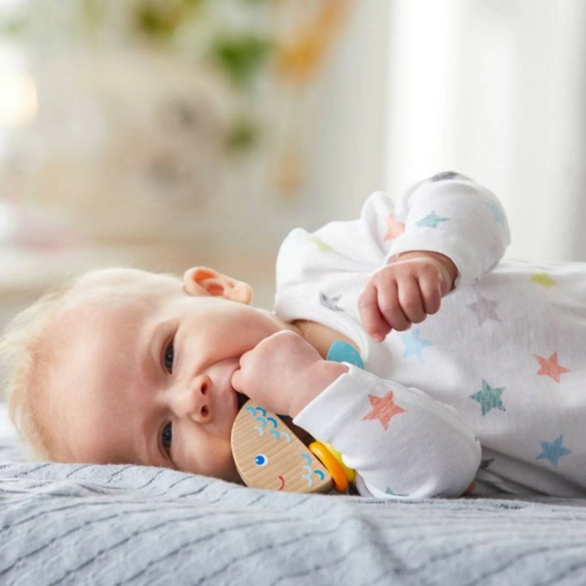 Baby lying on a bed with a teething ring, wearing a onesie with star patterns.