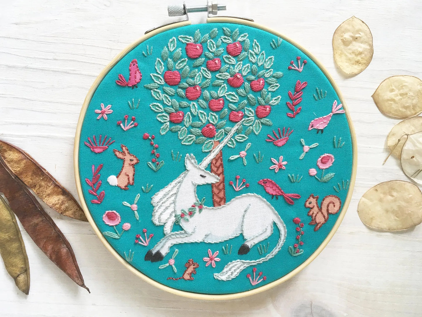 Embroidery hoop with a unicorn design on a teal background, surrounded by natural elements.