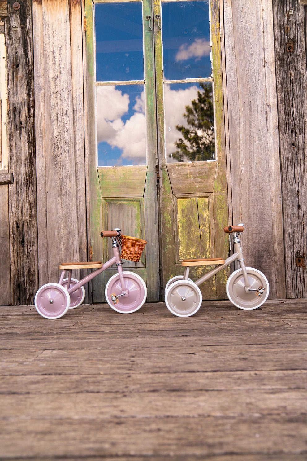 Two children's tricycles on a wooden floor with a rustic wooden wall and open door in the background.