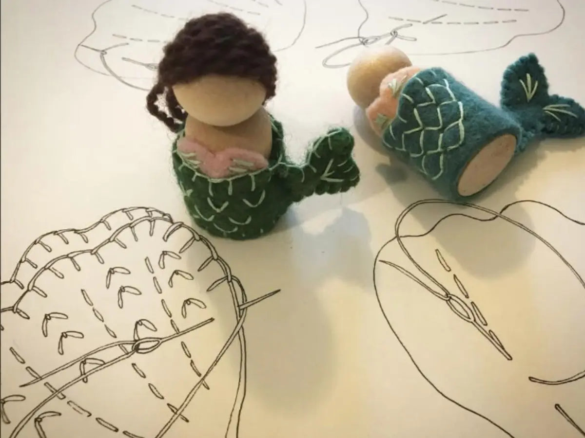 Two small mermaid peg dolls with green felt on a surface with hand-drawn lines and shapes. 