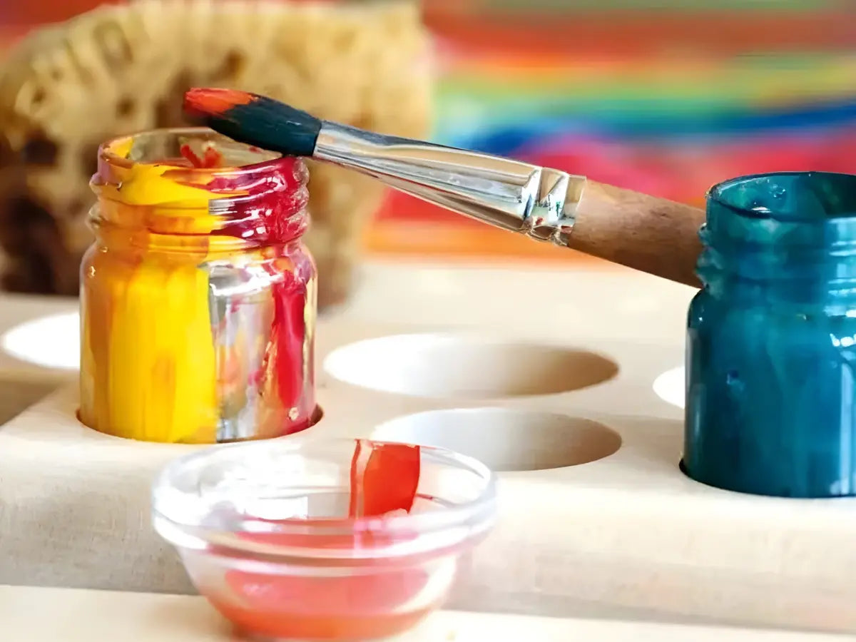 Paintbrush with red paint touching a jar of yellow paint, surrounded by other paint containers on a wooden surface.