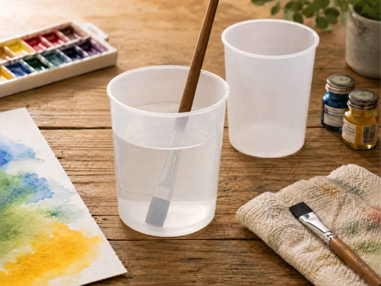 Two translucent watercolor water pots on a rustic wooden table with long-handled flat watercolor brushes, paint jars, cloth, and watercolor paper