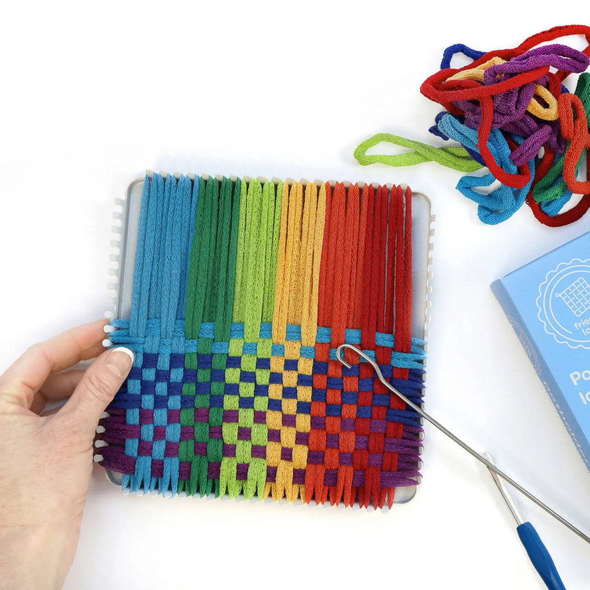 Hand weaving rainbow cotton loops over and under on a metal potholder loom with a weaving hook