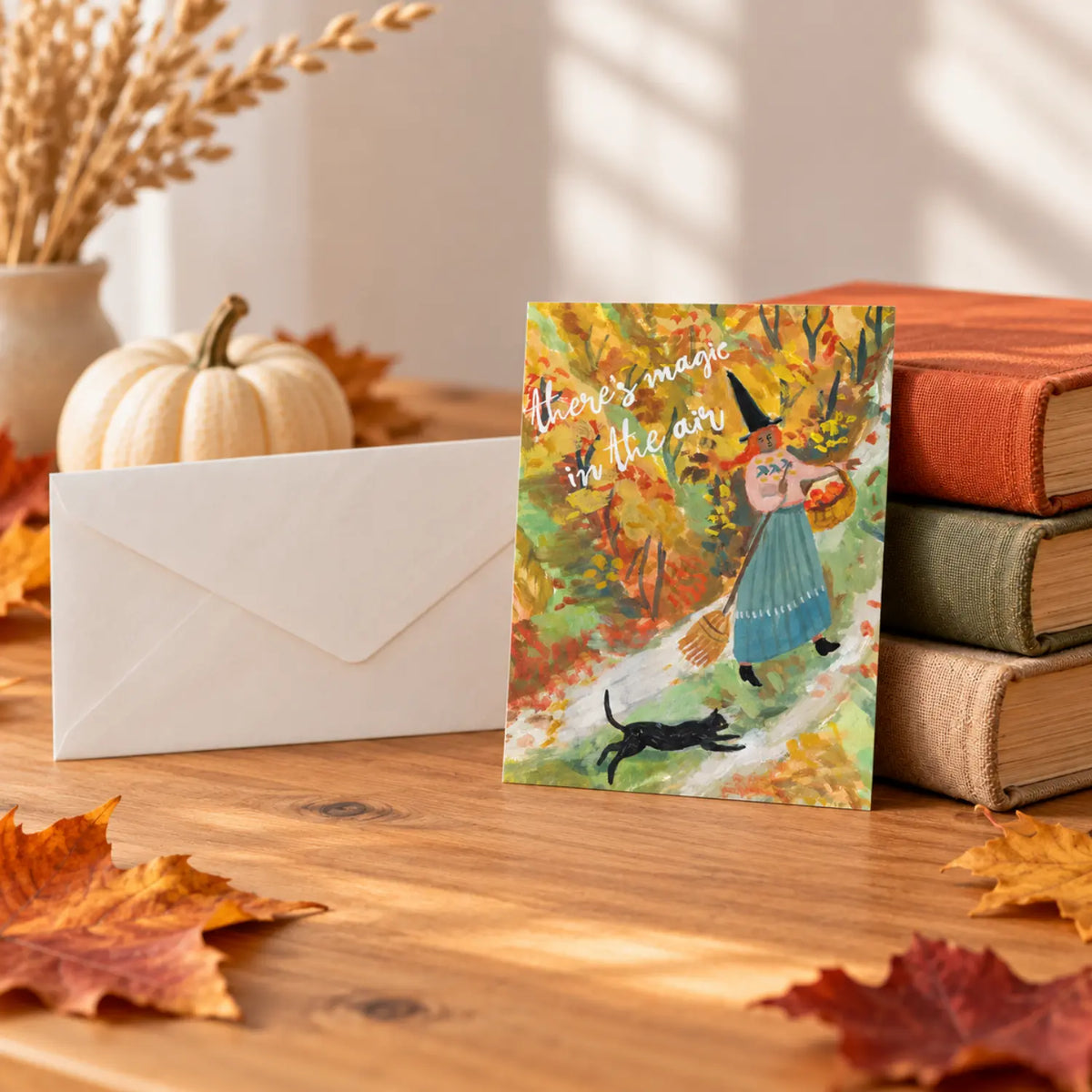 There’s Magic in the Air Halloween postcard by Ingrid Press displayed with autumn books, leaves, a pale pumpkin, and an envelope.