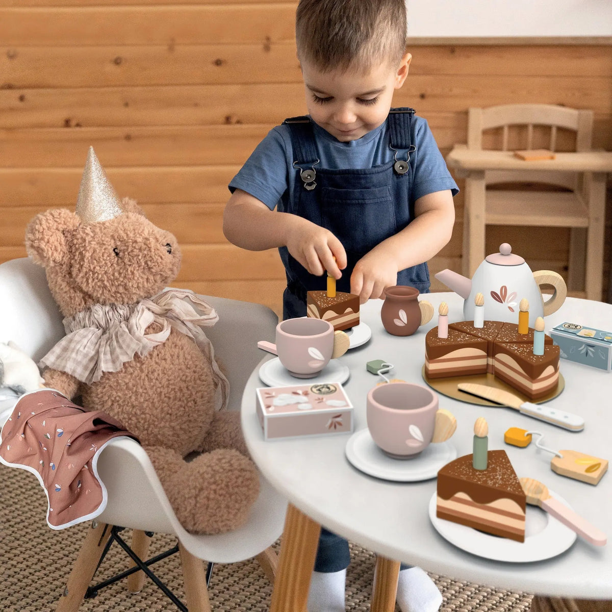 Child hosting a pretend birthday tea party with a wooden tea set, chocolate cake, candles, teapot, cups, and teddy bear.