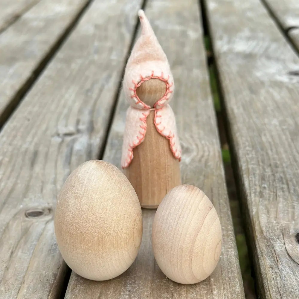Wooden gnome with pink outfit standing between two wooden eggs on a wooden surface