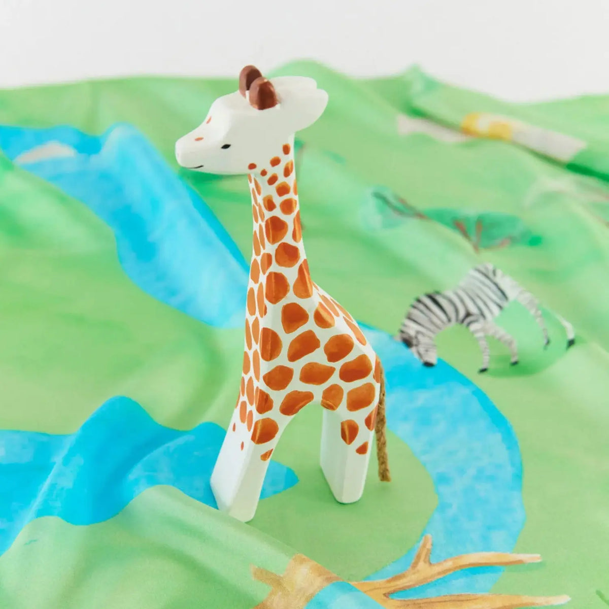 Wooden giraffe toy standing on a green and blue river playsilk with a wooden zebra in the background