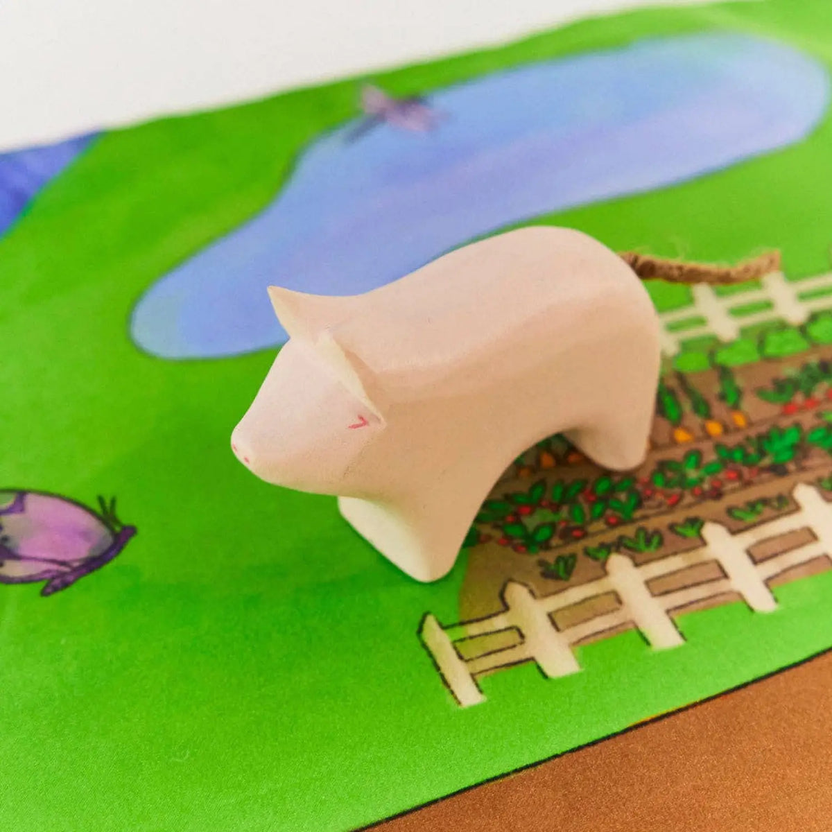 Wooden pig toy beside a fenced vegetable garden and pond on a farm play mat