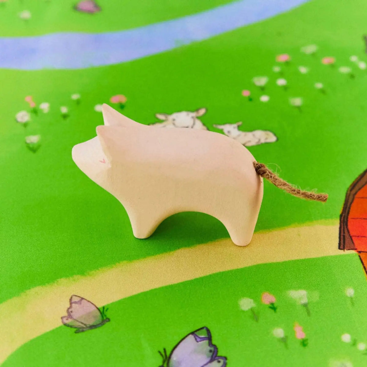 Wooden pig toy in a flowered farm meadow with sheep, a path, and a red barn on a play mat
