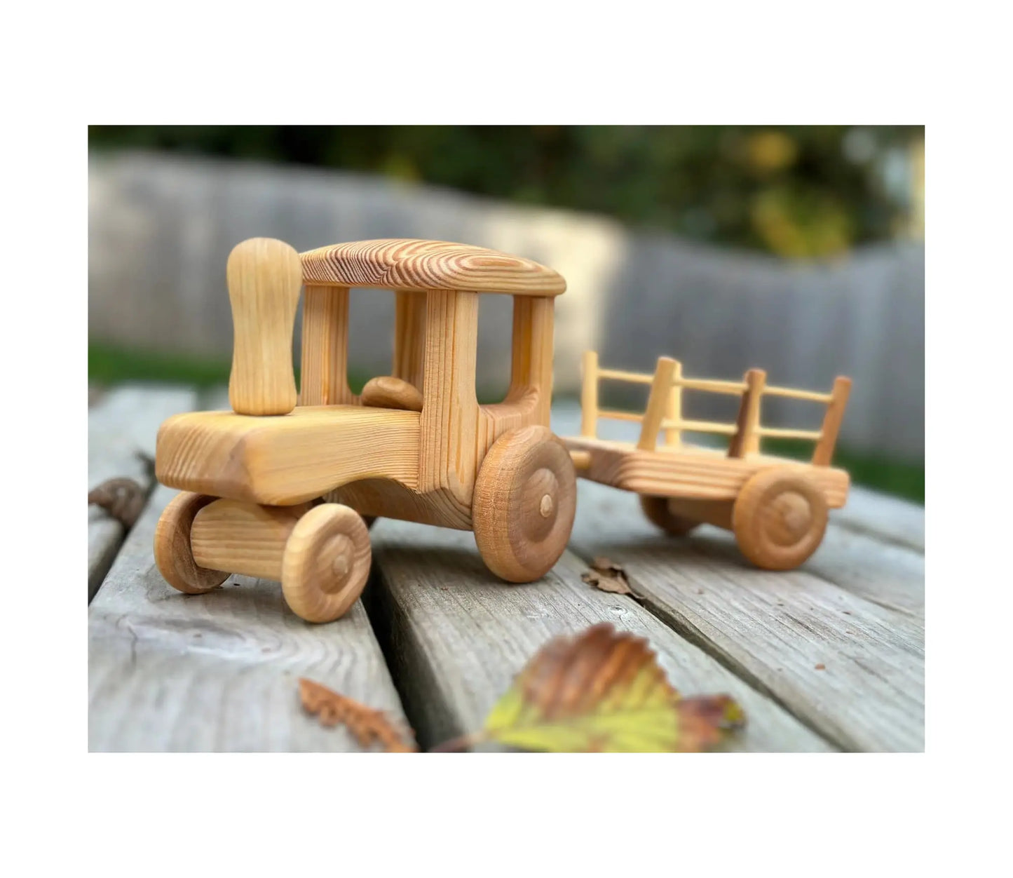 Wooden tractor with trailer shown from front angle outdoors on wooden surface with autumn leaves