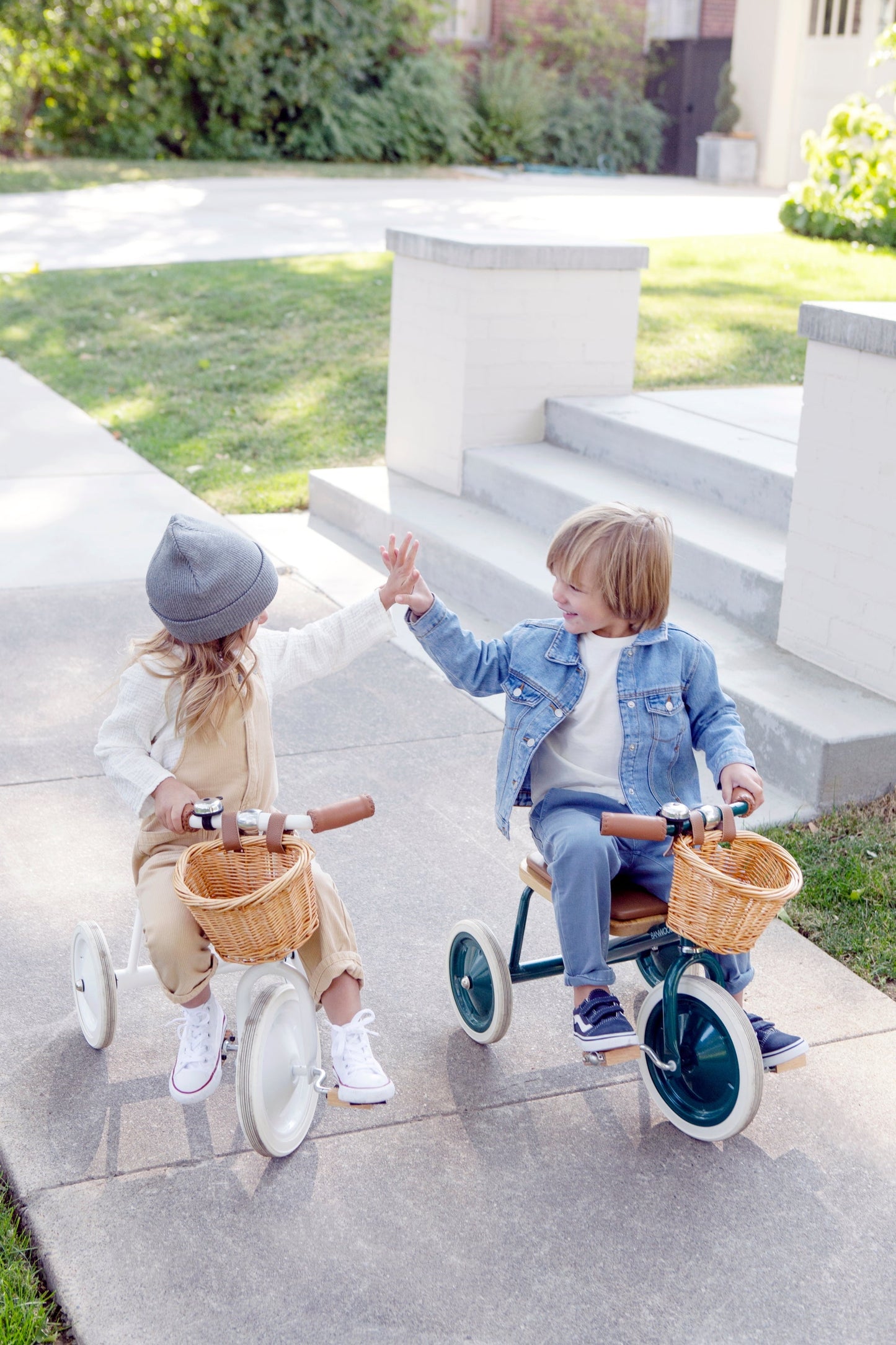 Two kids riding Banwood vintage trike toddler tricycles with basket on sidewalk