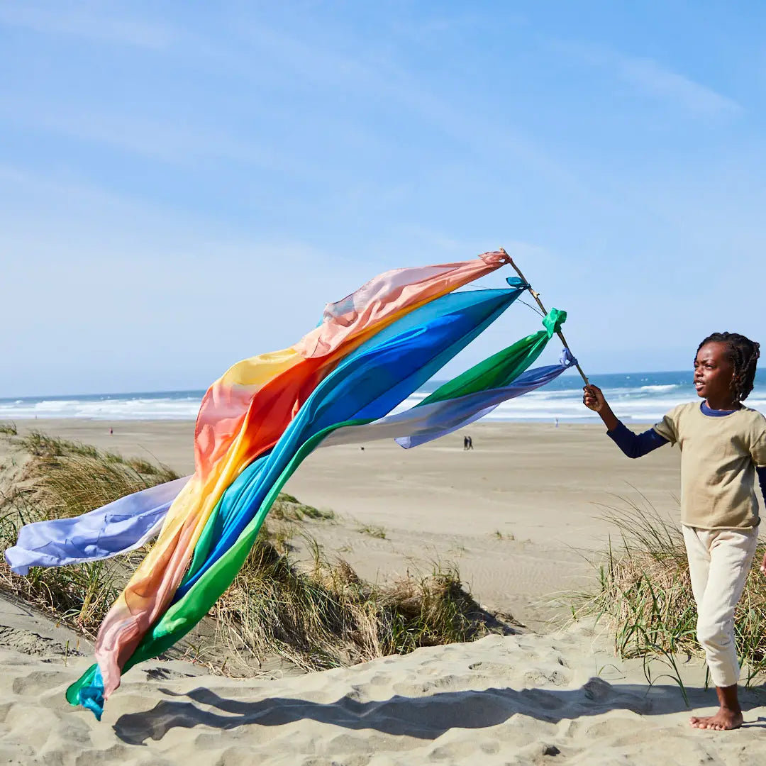 Girl walking on beach holding long stick with multiple colorful giant playsilks tied on blowing in wind