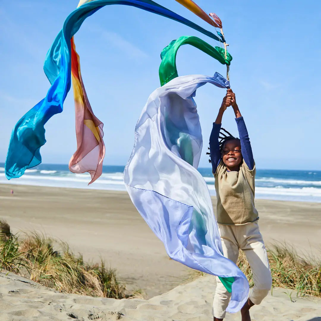Girl jumping on beach holding long stick with multiple colorful giant playsilks tied on blowing in wind