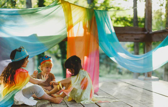 Three children playing under a rainbow giant playsilk in a treehouse