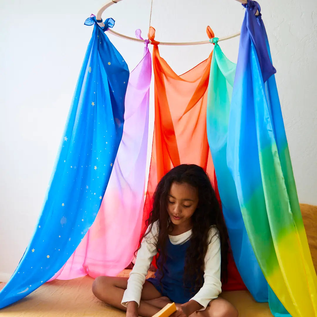 Girl sitting under several giant playsilks clipped to a ring hung from the ceiling